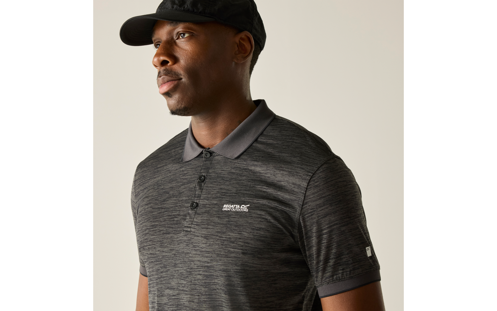 Borst-opwaarts portret van een persoon met een zwarte pet en donkergrijs poloshirt met klein logo, kijkt naar links tegen een lichte achtergrond