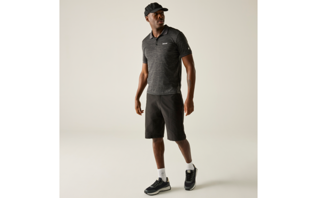 Persoon staand, licht gedraaid voor een lichte achtergrond, draagt een donker poloshirt, donkere knielange shorts, een zwarte pet, witte sokken en zwarte sneakers, kijkt opzij