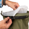 Deux mains : l'une tient le bord supérieur d'un vêtement imperméable olive à finition noire, l'autre introduit un sachet plastique blanc translucide