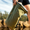 Main tenant un sac roulé vert olive avec boucles et textes imprimés ; rochers et arbres flous en arrière-plan ; partie d'une cuisse en vêtement noir visible