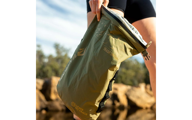 Main tenant un sac roulé vert olive avec boucles et textes imprimés ; rochers et arbres flous en arrière-plan ; partie d'une cuisse en vêtement noir visible