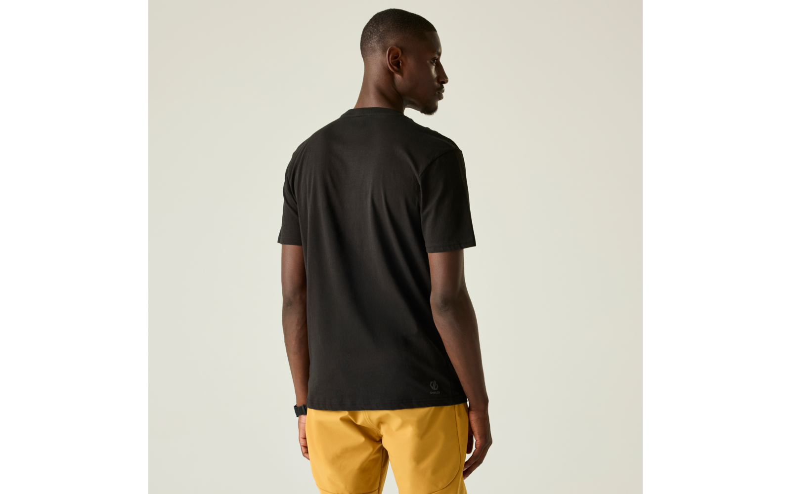 Person von hinten, trägt schwarzes kurzärmeliges T-Shirt und gelbe kurze Hose, steht vor hellem Hintergrund, Kopf nach rechts gedreht