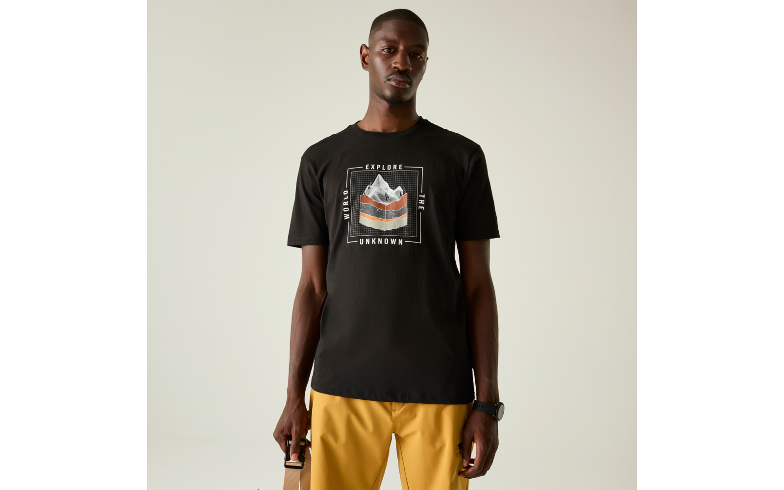 Ein Mann steht frontal vor hellem, einfarbigem Hintergrund. Er trägt ein schwarzes T-Shirt mit Berggrafik und dem weißen Schriftzug "EXPLORE THE UNKNOWN" sowie gelbe Hosen. Er hält einen beigen Taschenhenkel in einer Hand und trägt eine schwarze Armb