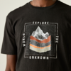 Oberkörper einer Person in schwarzem T‑Shirt mit quadratischer Grafik: Rasterhintergrund, weiße Bergsilhouette, drei horizontale Schichten in Orange, Dunkelgrau und Beige; Text 'EXPLORE' oben, 'WORLD' links, 'THE' rechts, 'UNKNOWN' unten