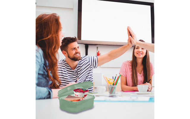 Drei Personen an einem Tisch. Linke Person mit roten Haaren im Profil. Mittlere Person mit gestreiftem Shirt hebt die Hand; eine Hand von rechts berührt sie. Rechte Person in rosa T‑Shirt lächelnd. Auf dem Tisch offene grüne Brotdose mit Sandwich und