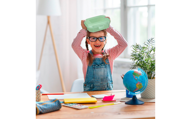 Kind mit Brille und Zöpfen hält eine grüne Brotdose über dem Kopf, sitzt an einem Tisch mit Schreibutensilien, Heften und einem Globus