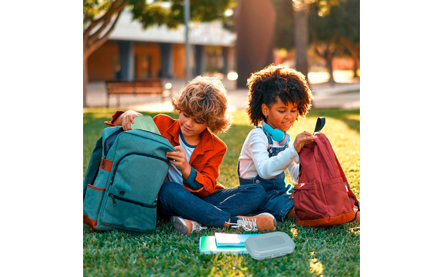 Zwei Kinder sitzen auf dem Gras neben zwei Rucksäcken; eines trägt Kopfhörer um den Hals. Vor ihnen liegen Hefte und eine Brotdose, im Hintergrund sind Bäume und Bänke zu sehen