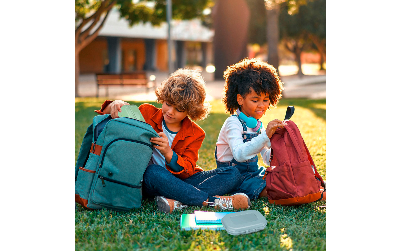 Zwei Kinder sitzen auf dem Gras neben zwei Rucksäcken; eines trägt Kopfhörer um den Hals. Vor ihnen liegen Hefte und eine Brotdose, im Hintergrund sind Bäume und Bänke zu sehen