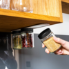 Eine Hand hält ein kleines Glas mit gelben Senfkörnern, entnommen von einem Set aus drei kleinen Glasgefäßen mit schwarzen Deckeln, die unter einem Holzregal befestigt sind; die anderen Gläser enthalten getrocknete rote Chilischoten und Lorbeerblätte