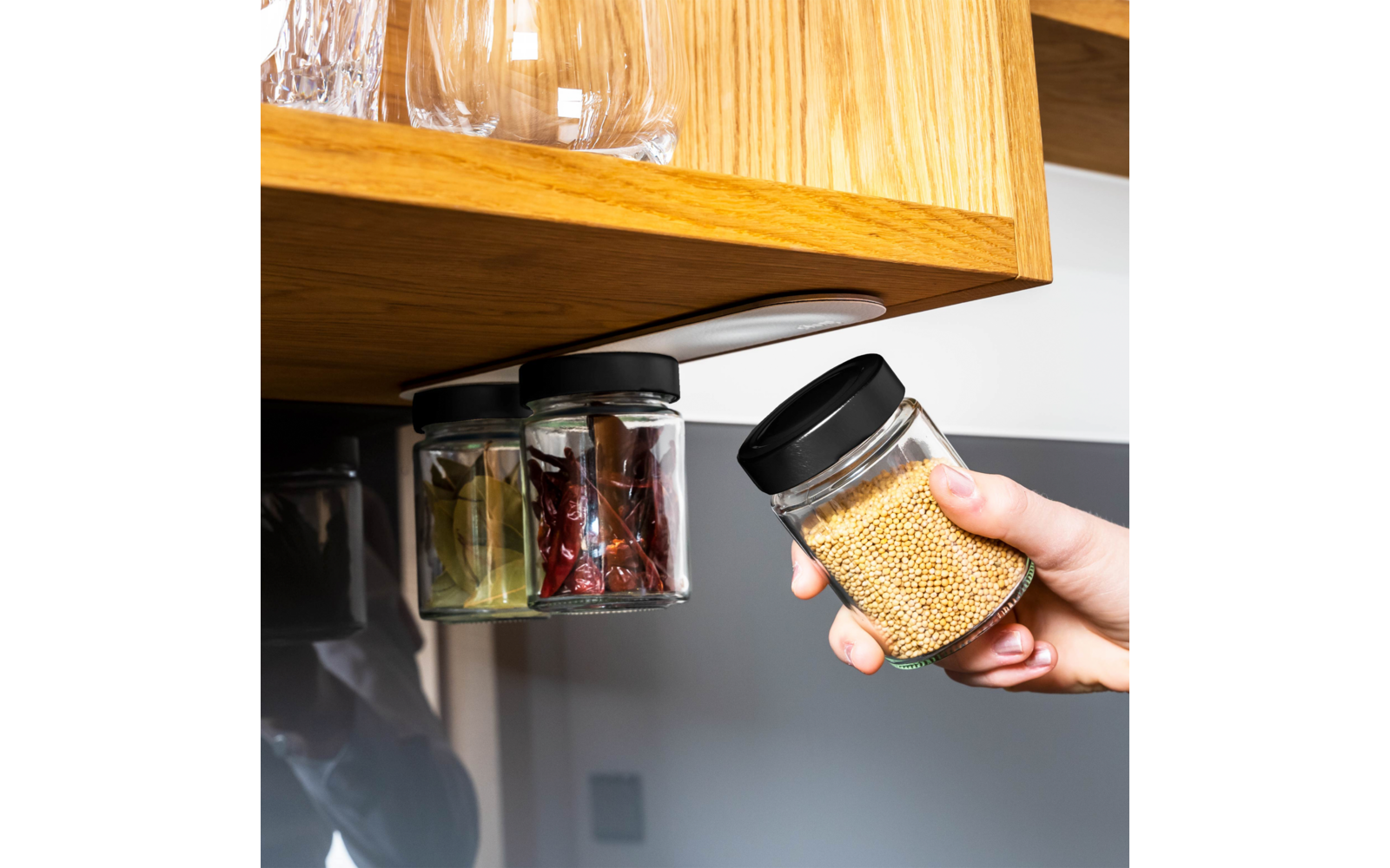 Eine Hand hält ein kleines Glas mit gelben Senfkörnern, entnommen von einem Set aus drei kleinen Glasgefäßen mit schwarzen Deckeln, die unter einem Holzregal befestigt sind; die anderen Gläser enthalten getrocknete rote Chilischoten und Lorbeerblätte