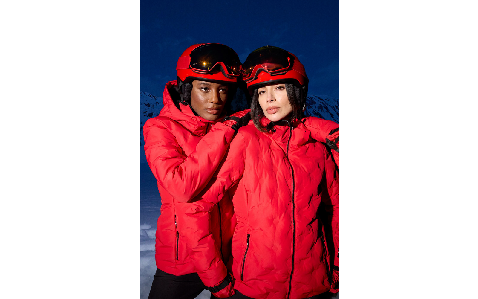 Ein Mann und eine Frau tragen rote Skianzüge und Helme mit Skibrillen vor einem dunklen Hintergrund