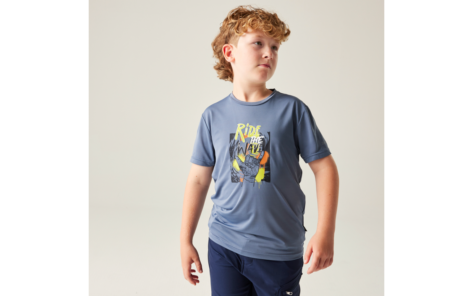 Junge mit lockigem blondem Haar trägt blaues T‑Shirt mit Aufdruck 'Ride the Wave' und dunkelblaue Shorts, schaut leicht nach rechts