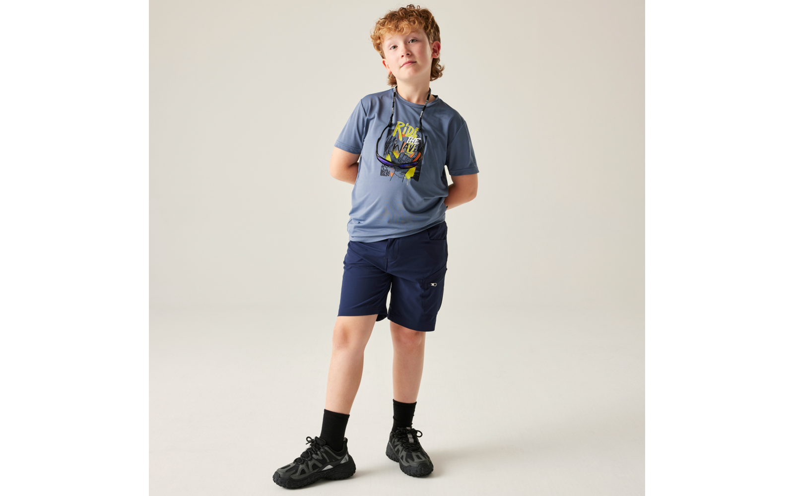 Stehendes Kind mit lockigem rötlich-blondem Haar, blauem T-Shirt mit Print, dunkelblauen Shorts, schwarzen Socken und Turnschuhen, Hände hinter dem Rücken, heller Hintergrund