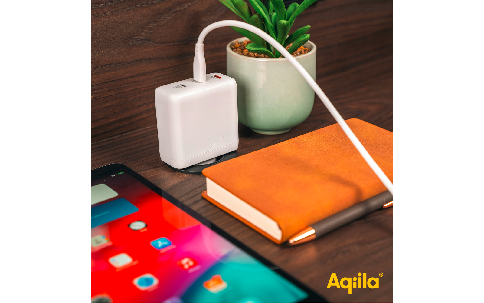 Ladekabel, das an einem weißen Ladegerät angeschlossen ist, liegt auf einem Holztisch neben einem orangefarbenen Notizbuch, einem Smartphone und einem Blumentopf mit einer grünen Pflanze