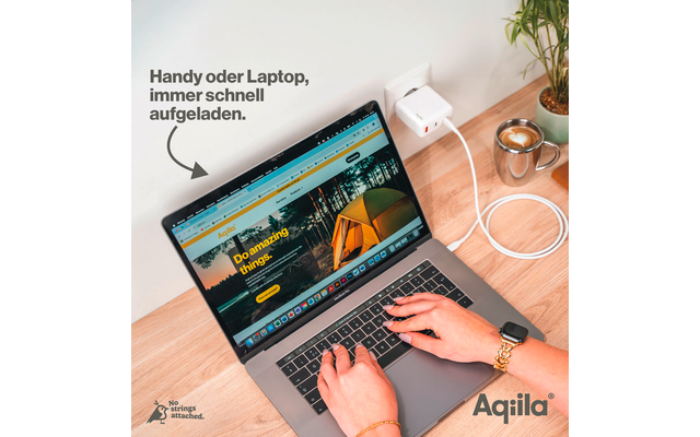 Laptop auf Tisch mit geöffnetem Bildschirm, Hände auf Tastatur, daneben Smartphone, Pflanze und Kabel