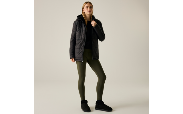 Persona in piedi davanti a uno sfondo neutro chiaro, indossa una giacca imbottita nera con cappuccio, un top nero sotto, leggings verde oliva, stivaletti neri con bordo in pelliccia, una mano tiene la giacca al colletto, capelli biondi fino alle spalle