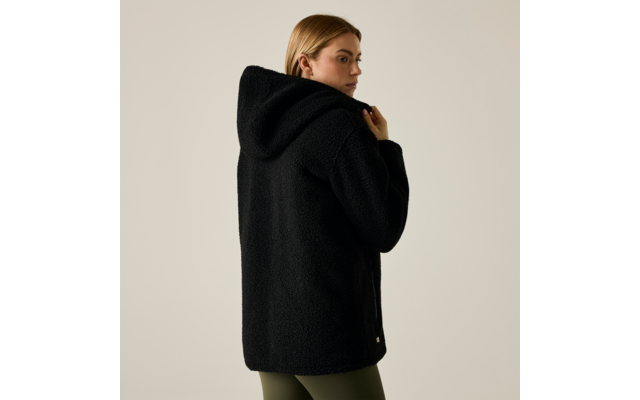 Persona con capelli biondi fino alle spalle, vista di schiena e di profilo, indossa una giacca teddy nera con cappuccio e leggings verde oliva, sfondo chiaro uniforme