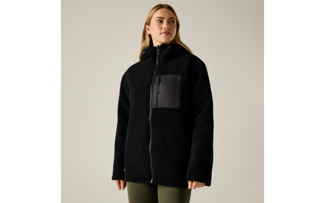 Persona che indossa una giacca in pile nera con cerniera e tasca rettangolare sul petto, pantaloni verde oliva, in piedi davanti a uno sfondo chiaro uniforme, sguardo leggermente verso destra
