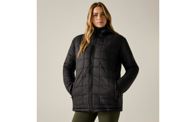 Persona con lunghi capelli biondi, in piedi frontalmente, indossa una giacca imbottita nera trapuntata con cerniera e cappuccio, mani nelle tasche, leggings verde oliva su sfondo chiaro uniforme