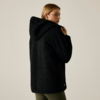Persona con capelli biondi fino alle spalle, vista di schiena e di profilo, indossa una giacca teddy nera con cappuccio e leggings verde oliva, sfondo chiaro uniforme