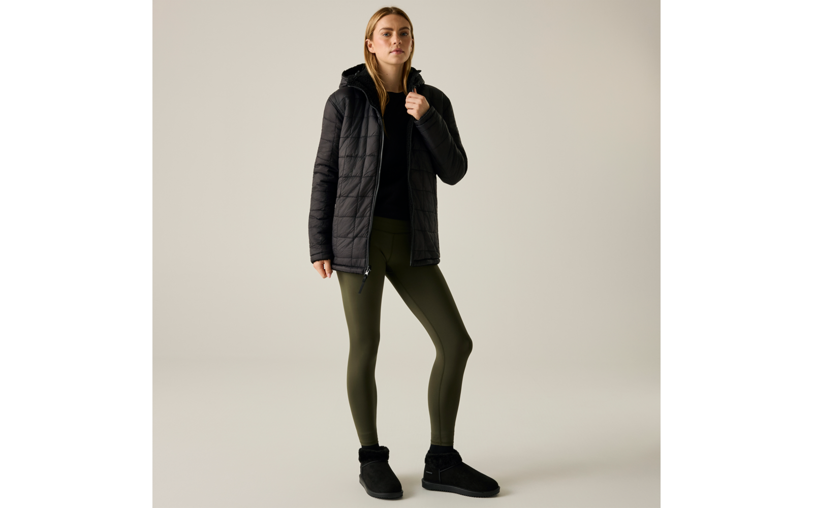 Persona in piedi davanti a uno sfondo neutro chiaro, indossa una giacca imbottita nera con cappuccio, un top nero sotto, leggings verde oliva, stivaletti neri con bordo in pelliccia, una mano tiene la giacca al colletto, capelli biondi fino alle spalle