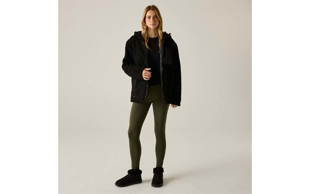Donna in piedi di fronte a uno sfondo chiaro, indossa una giacca in pile nera con cappuccio e zip, una maglia nera, leggings verde oliva e stivaletti neri; una mano tiene la zip