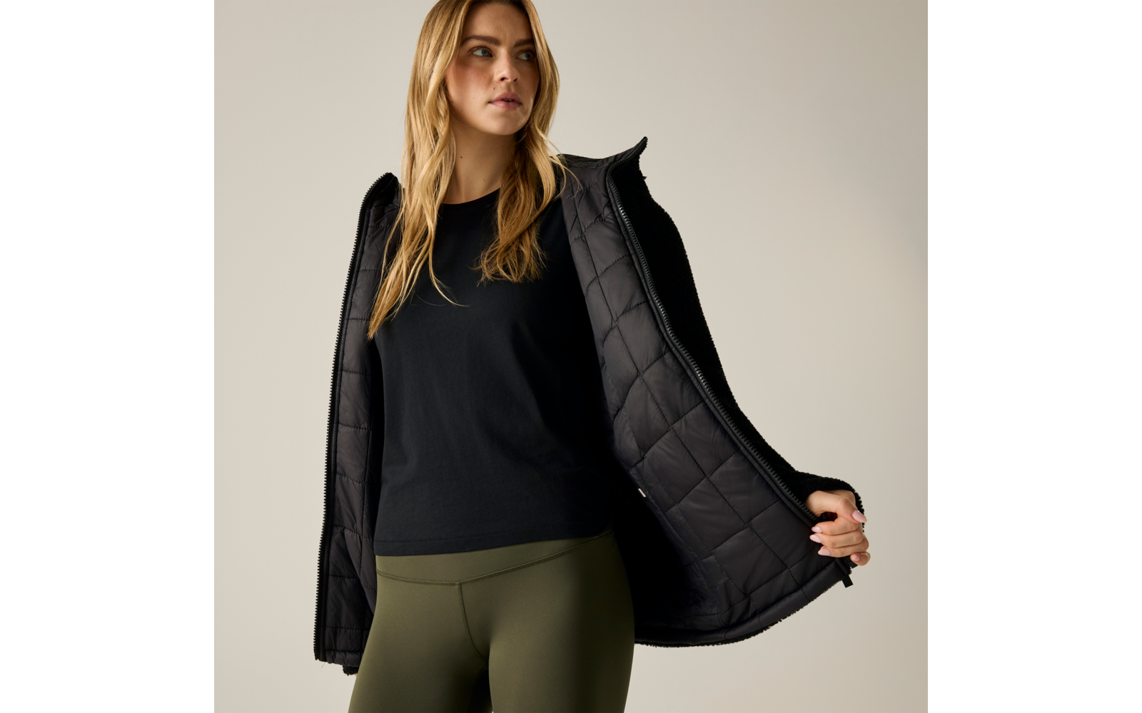 Persona con lunghi capelli biondi che indossa una maglia nera a maniche lunghe, una giacca imbottita nera aperta e leggings verde oliva su sfondo neutro