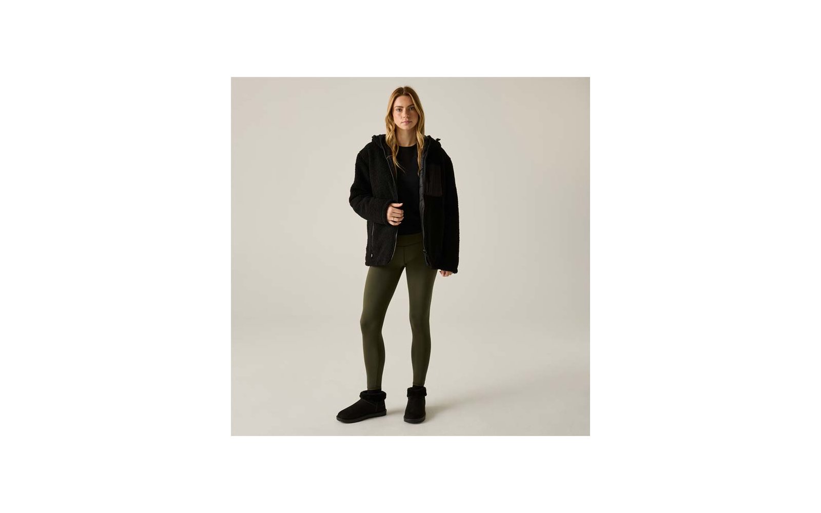 Donna in piedi di fronte a uno sfondo chiaro, indossa una giacca in pile nera con cappuccio e zip, una maglia nera, leggings verde oliva e stivaletti neri; una mano tiene la zip