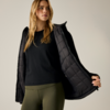 Persona con lunghi capelli biondi che indossa una maglia nera a maniche lunghe, una giacca imbottita nera aperta e leggings verde oliva su sfondo neutro