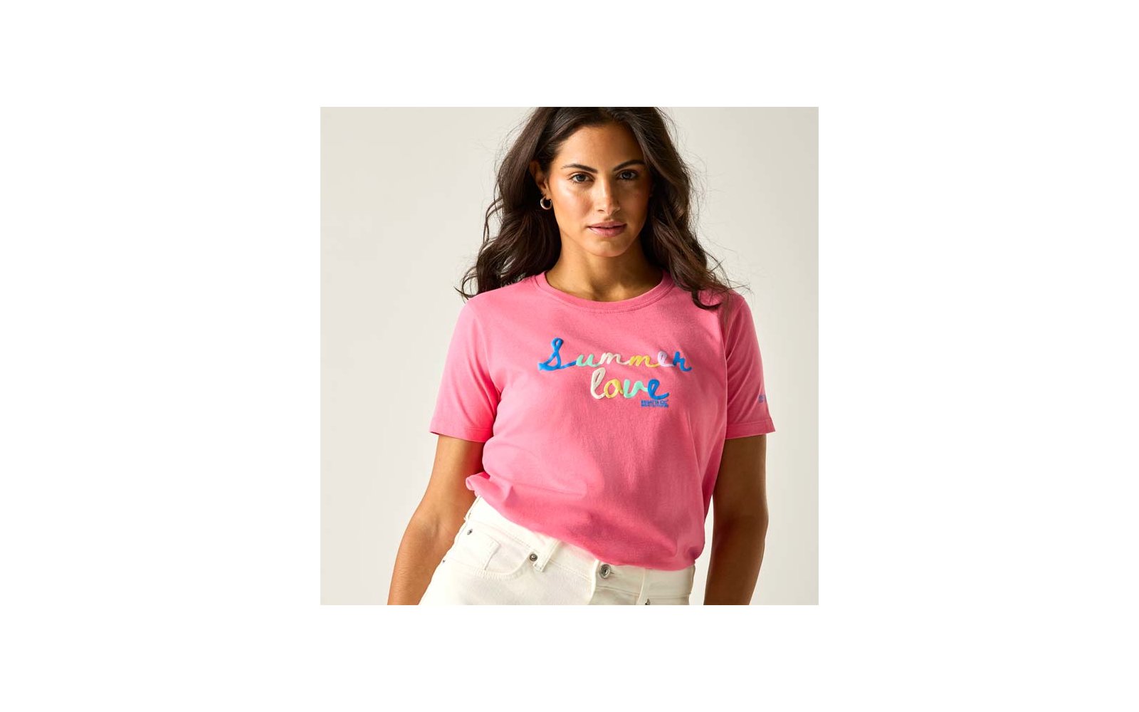 Person mit pinkem T-Shirt mit mehrfarbigem Schriftzug ‚Summer love‘, weiße Hose, sichtbar vom Oberkörper bis zu den Hüften, dunkle lange Haare, Creolen, heller Hintergrund