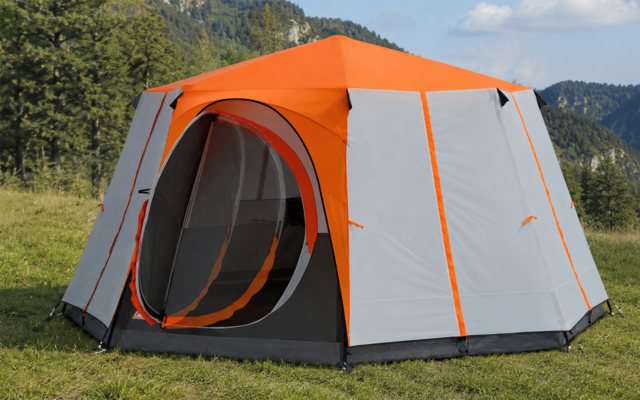 Orange and gray tent with a round open entrance on a grassy meadow; trees, mountains and blue sky in the background