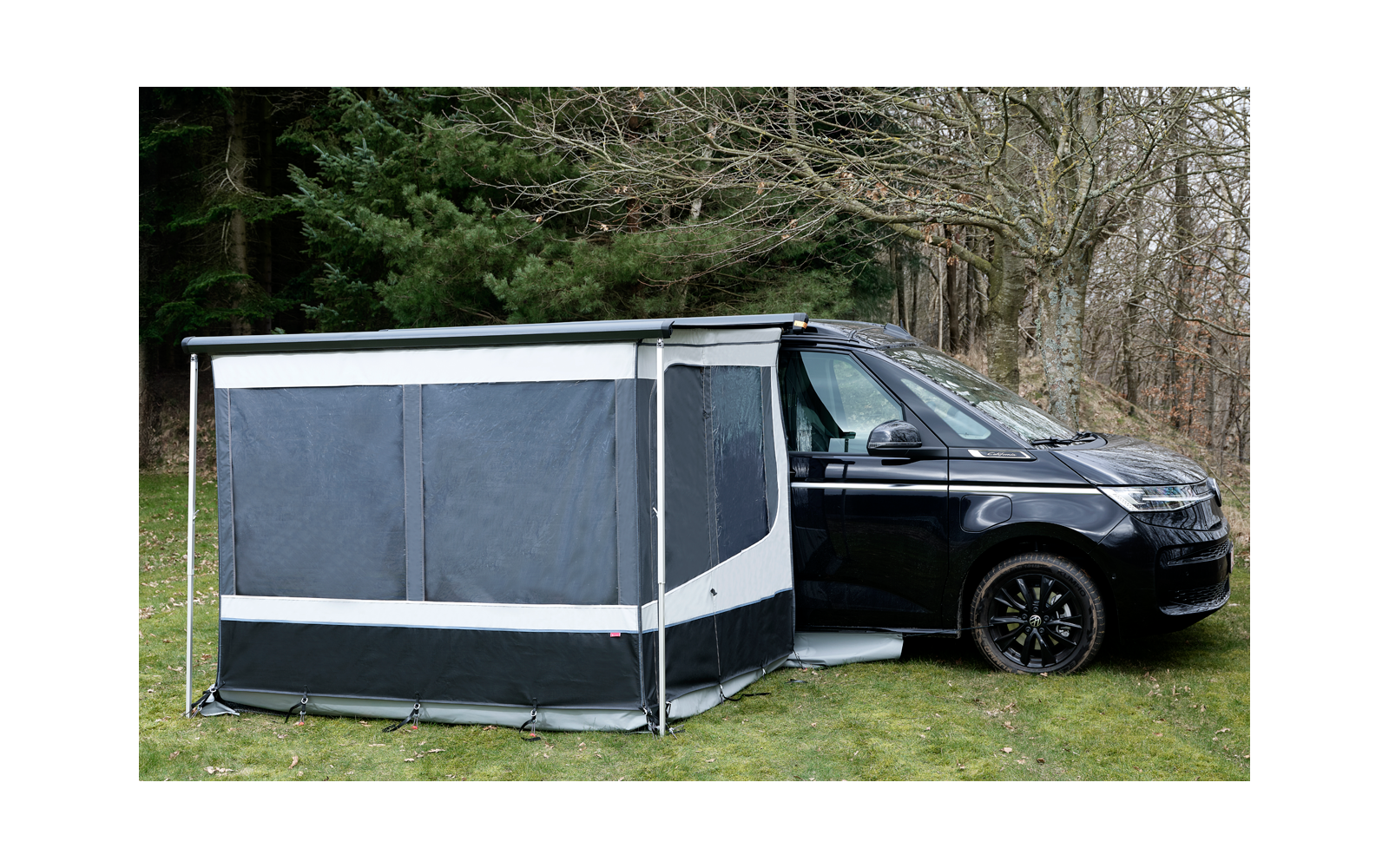 Schwarzer Campingbus auf Gras neben kahlen Bäumen, an der Seite ein grau-schwarzes Vorzelt mit geschlossenen Fenstern und weißen Streifen
