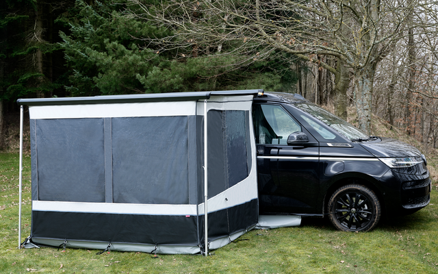 Schwarzer Campingbus auf Gras neben kahlen Bäumen, an der Seite ein grau-schwarzes Vorzelt mit geschlossenen Fenstern und weißen Streifen