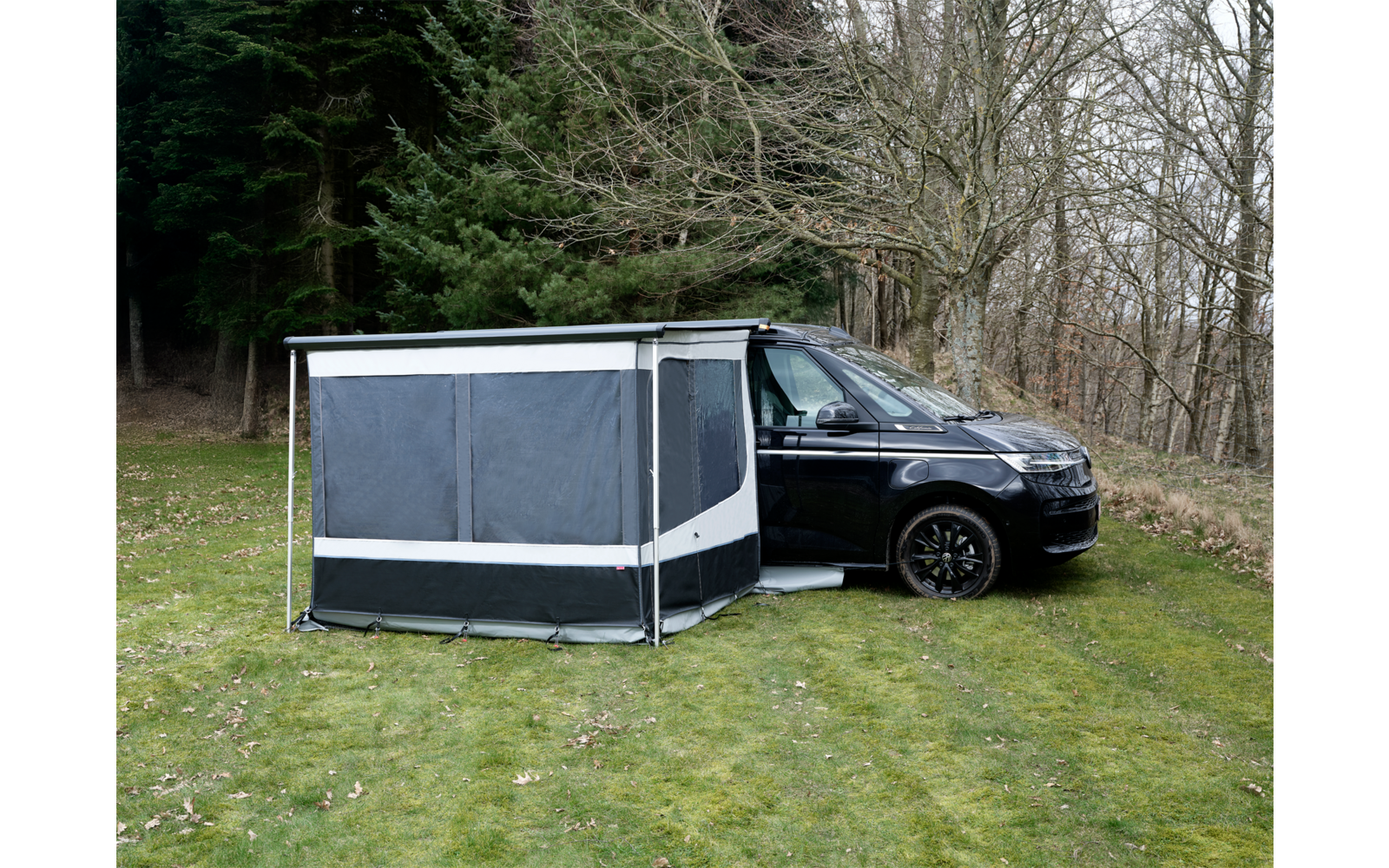 Schwarzer Campingbus auf Gras neben kahlen Bäumen, an der Seite ein grau-schwarzes Vorzelt mit geschlossenen Fenstern und weißen Streifen