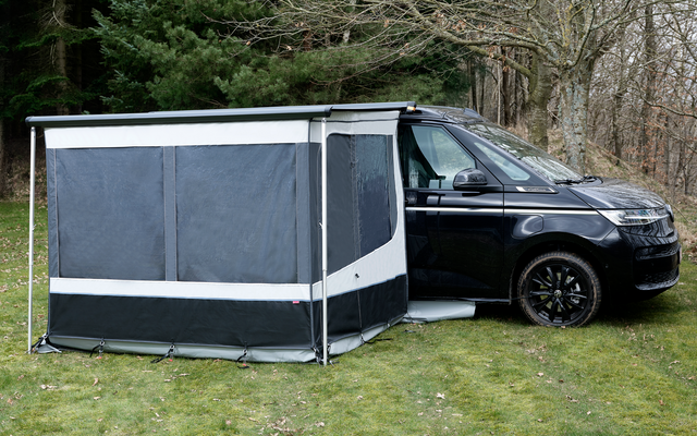 Furgoneta camper negra sobre césped junto a árboles sin hojas, con un toldo gris y negro fijado al lateral, paneles de ventanas cerrados y ribete blanco