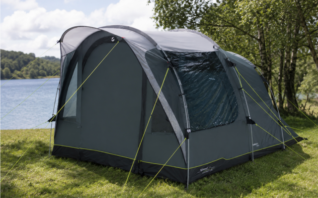 Groen-grijze tent met scheerlijnen op gras aan een oever, bomen rechts, meer en bewolkte lucht op de achtergrond