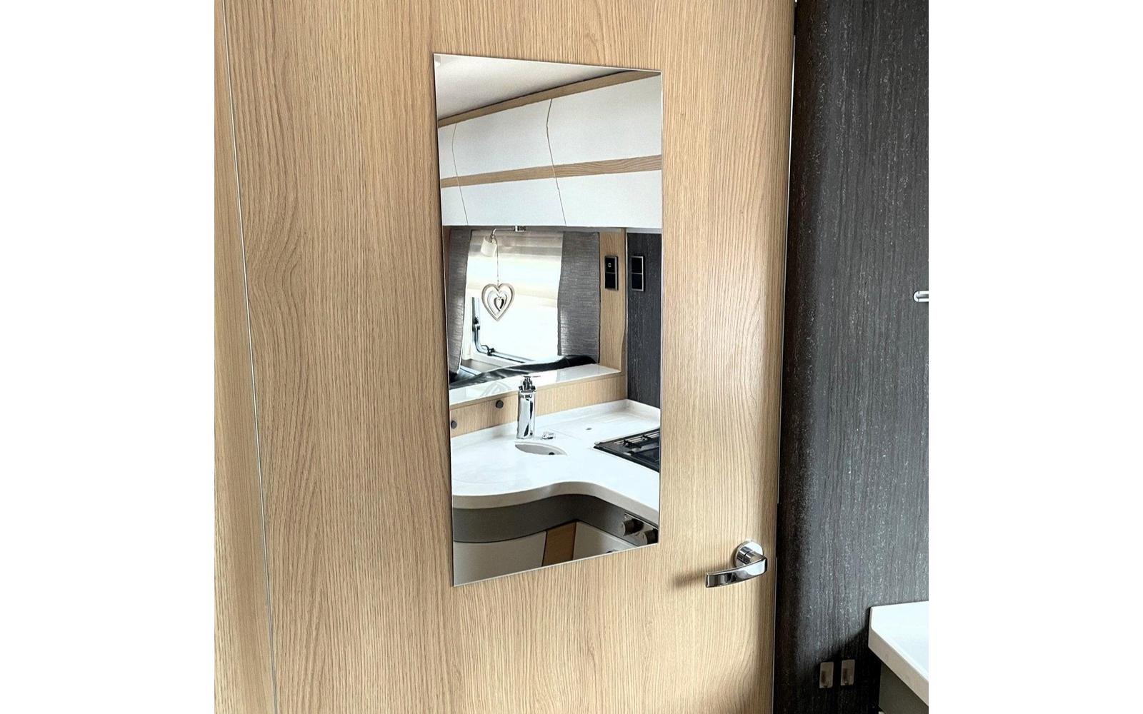 Rechteckiger Spiegel in Holztür, im Spiegel eine Küchenzeile mit weißer Arbeitsfläche, Spüle mit Armatur, Herd, Hängeschränken und Herz-Anhänger am Fenster