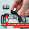 Main tenant un bouchon argenté au-dessus d’un petit bocal en verre rempli de granulés blancs ; flèche courbée rouge et bannières rouges portant le texte « FESTSITZENDE VERSCHLUSSKAPPE » et « Aroma bleibt lange erhalten », arrière‑plan flou avec d’autres récipients