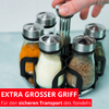 Main saisit un bocal en verre rond avec couvercle métallique dans un support noir; cinq bocaux en verre contenant des poudres colorées (jaune, blanc, brun, vert) sur fond bleu clair; bandeau rouge en bas avec texte blanc 'EXTRA GROSSER GRIFF Für den sicheren Transport des Rondells'