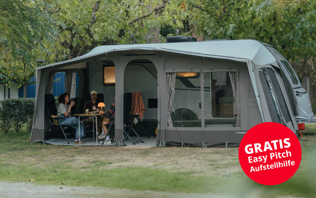 Large gray camping awning with multiple rectangular windows, dark base strip, curtains at the windows, curved roof, guy lines and ground pegs; red round sticker at lower right with white text
