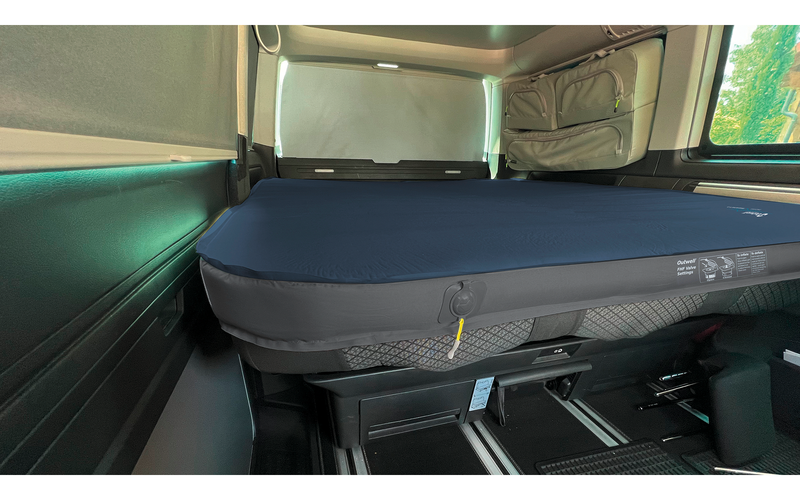 Blue inflatable mattress on a black sleeping platform inside a vehicle interior; gray fabric storage pocket on the side wall, window with partially closed blind, green foliage visible outside