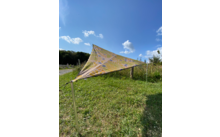 Dreieckiges, bunt gemustertes Sonnensegel auf einer grünen Wiese bei blauem Himmel mit einigen Wolken
