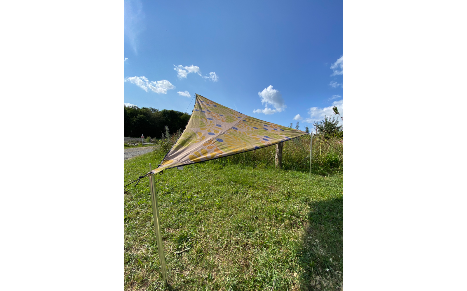 Dreieckiges, bunt gemustertes Sonnensegel auf einer grünen Wiese bei blauem Himmel mit einigen Wolken