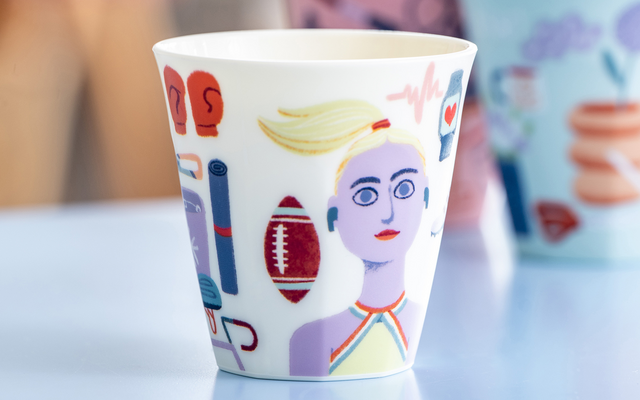 Tasse en céramique colorée au premier plan avec visage imprimé, ballon et symboles graphiques ; deux tasses peintes floues en arrière-plan