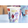 Tasse en céramique colorée au premier plan avec visage imprimé, ballon et symboles graphiques ; deux tasses peintes floues en arrière-plan