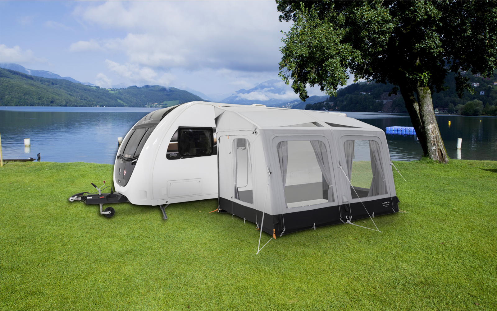 Ein Wohnwagen mit angebautem weißen Vorzelt steht auf einer grünen Wiese neben einem See