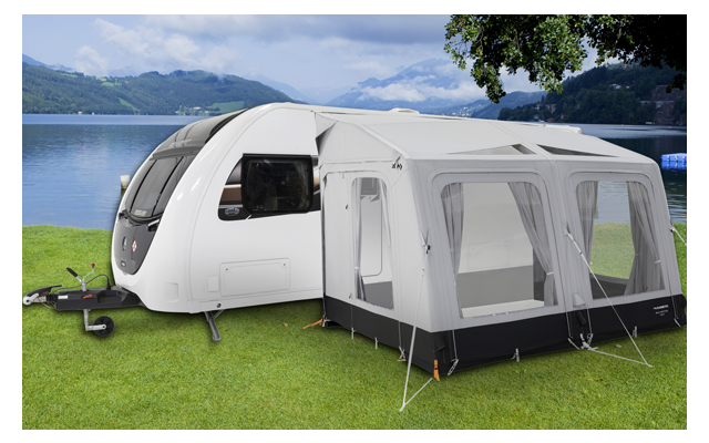 Ein Wohnwagen mit angebautem weißen Vorzelt steht auf einer grünen Wiese neben einem See