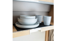 Gestapelte helle Schalen auf Tellern neben zwei Bechern auf einem Regal in einem Holzschrank