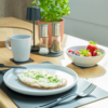 Tranche de pain complet avec tartinade blanche et herbes vertes hachées sur une assiette blanche; fourchette et couteau sur un set de table; tasse blanche; moulins à sel et poivre; bol avec fraises et fruits rouges mélangés; plante verte en pot sur une table en bois