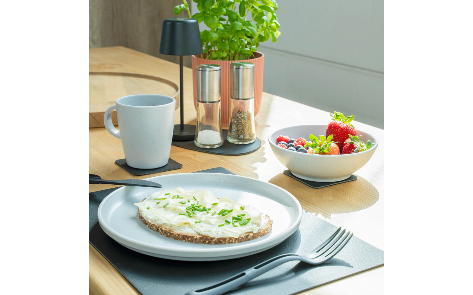 Tranche de pain complet avec tartinade blanche et herbes vertes hachées sur une assiette blanche; fourchette et couteau sur un set de table; tasse blanche; moulins à sel et poivre; bol avec fraises et fruits rouges mélangés; plante verte en pot sur une table en bois